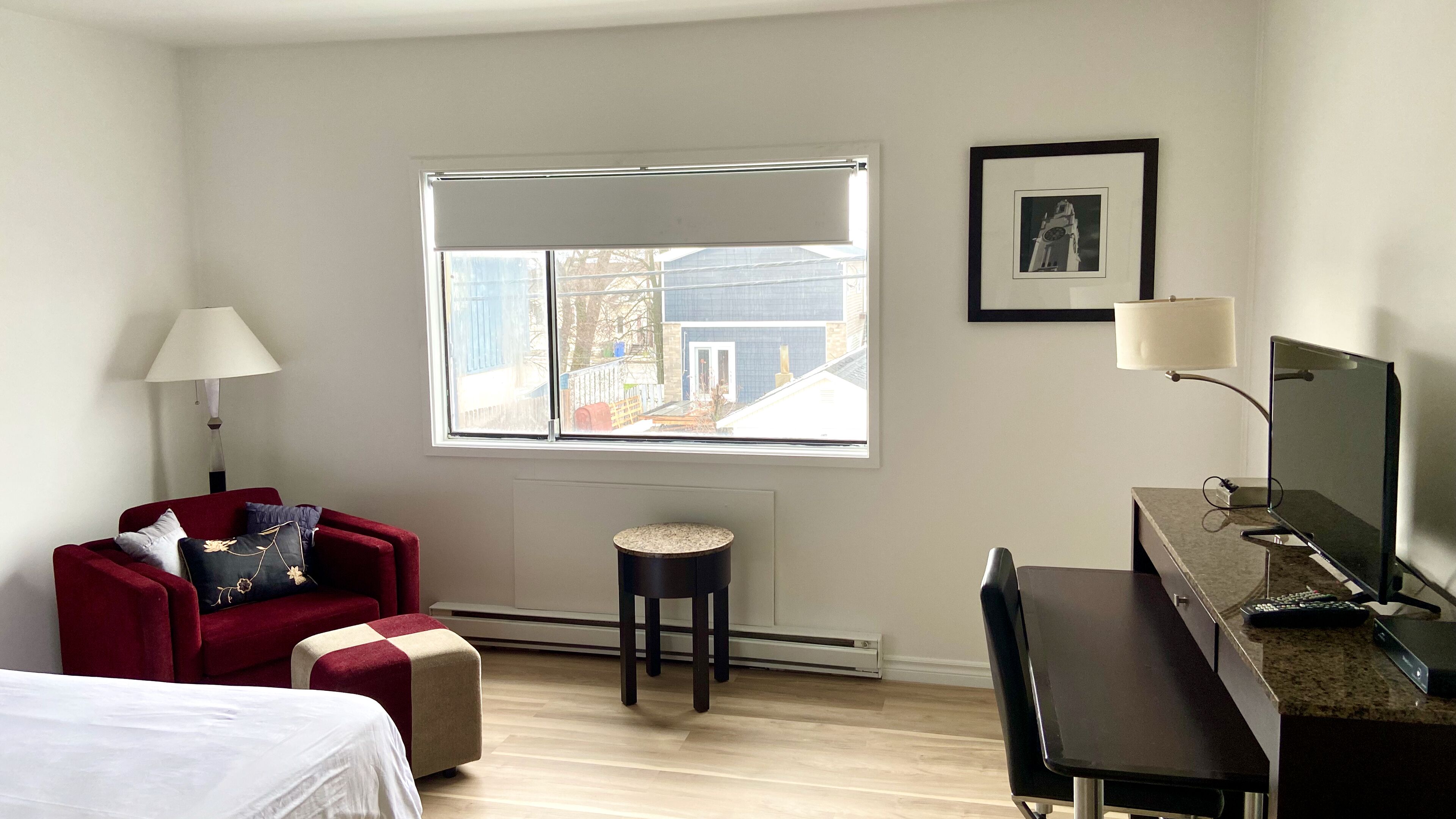 Una habitación de hotel con cama, un sillón rojo, un taburete pequeño, un escritorio con computadora y una ventana con vista a edificios.