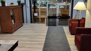 Zona de recepción con un mueble de madera, sillas rojas, suelo alfombrado y una puerta de cristal que da al exterior.
