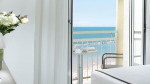 A hotel room with a balcony overlooking the sea, a bed, a small table with a cup and a book, and a vase with flowers.