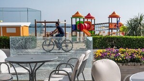 An outdoor seating area with tables and chairs, a playground with colorful play structures, and a person riding a bicycle.