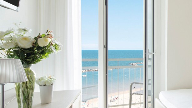 A hotel room with a balcony overlooking the sea, a bed, a desk with a lamp, and a vase with flowers.