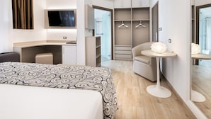A modern hotel room with a bed, desk, chair, wardrobe, and a round table with a lamp.