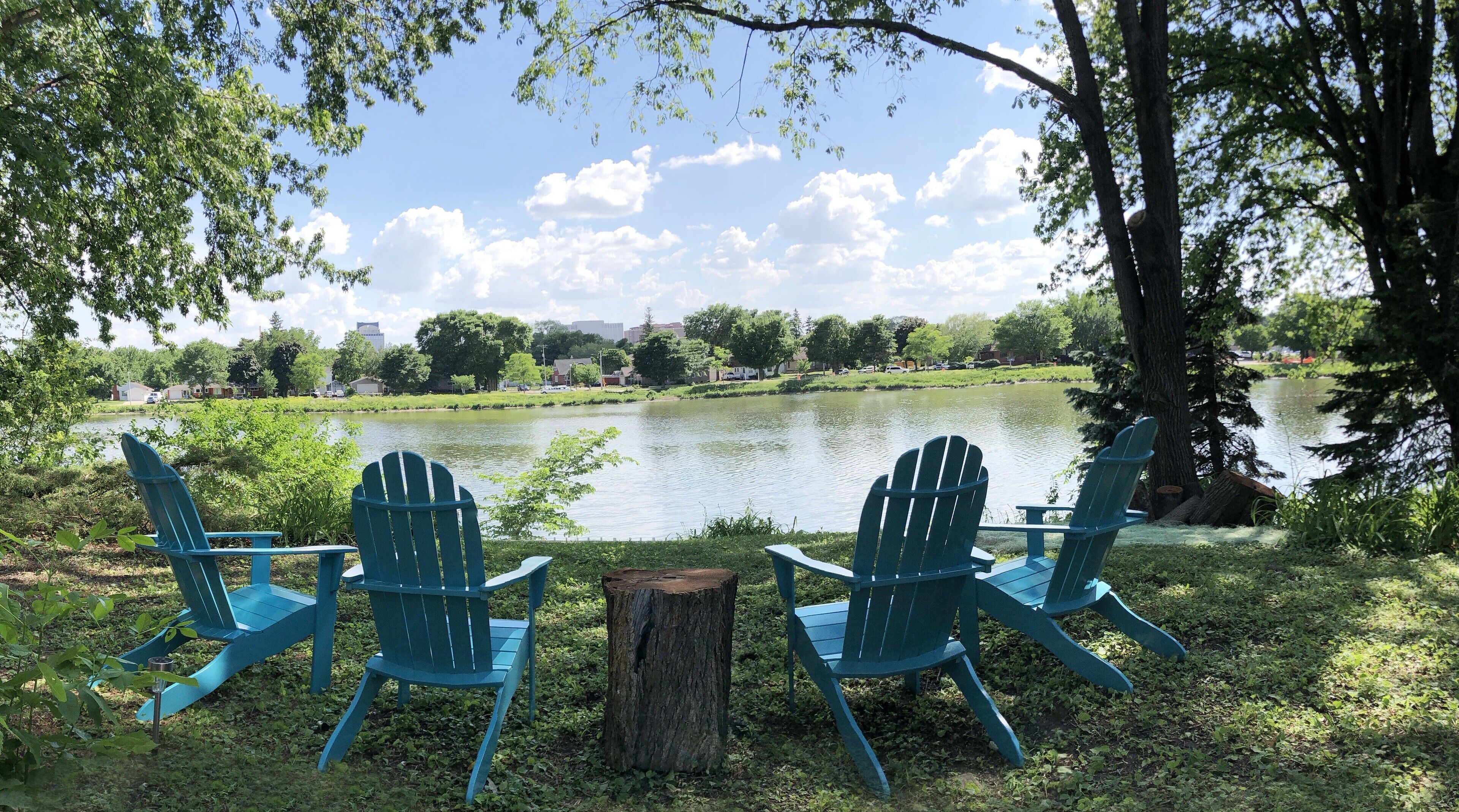 Trois fauteuils Adirondack sont placés face à un lac, offrant une vue sur un parc et des bâtiments situés au loin.