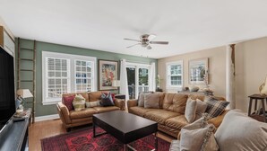 A living room with a brown leather sofa, a black coffee table, and a red patterned rug. There are two windows with white shutters, a ceiling fan, and a ladder against the wall.