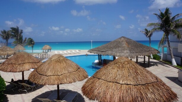 A pool area with thatched umbrellas, lounge chairs, and a view of the ocean. - New Beachfront PH at Brisas By The Spot (Cancun)