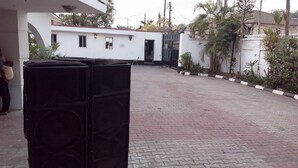 Un patio empedrado con un sistema de sonido negro, un edificio blanco con varias ventanas y una entrada con rejas.