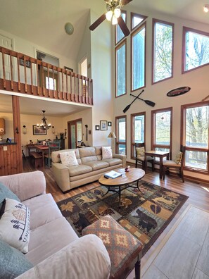 A spacious living area with a high ceiling, large windows, a wooden staircase, and a cozy seating arrangement.