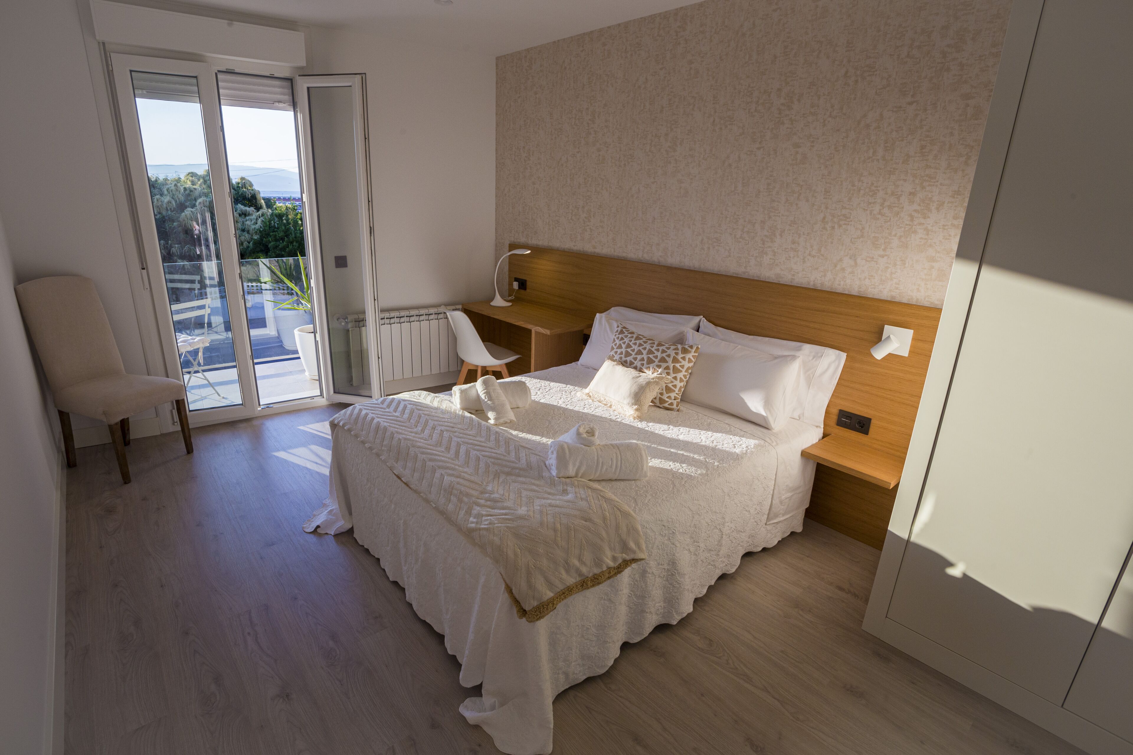 Une chambre avec un grand lit, une tête de lit en bois, un bureau avec une chaise et une vue sur l’extérieur, accessible par une porte-fenêtre coulissante.