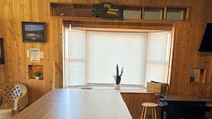 A wooden-paneled room with a large table, a window with blinds, a small stool, and a Pac-Man arcade machine. - Stay In Ohiopyle, Hot tub & Pool Table - Closest to the GAP trail (Ohiopyle)