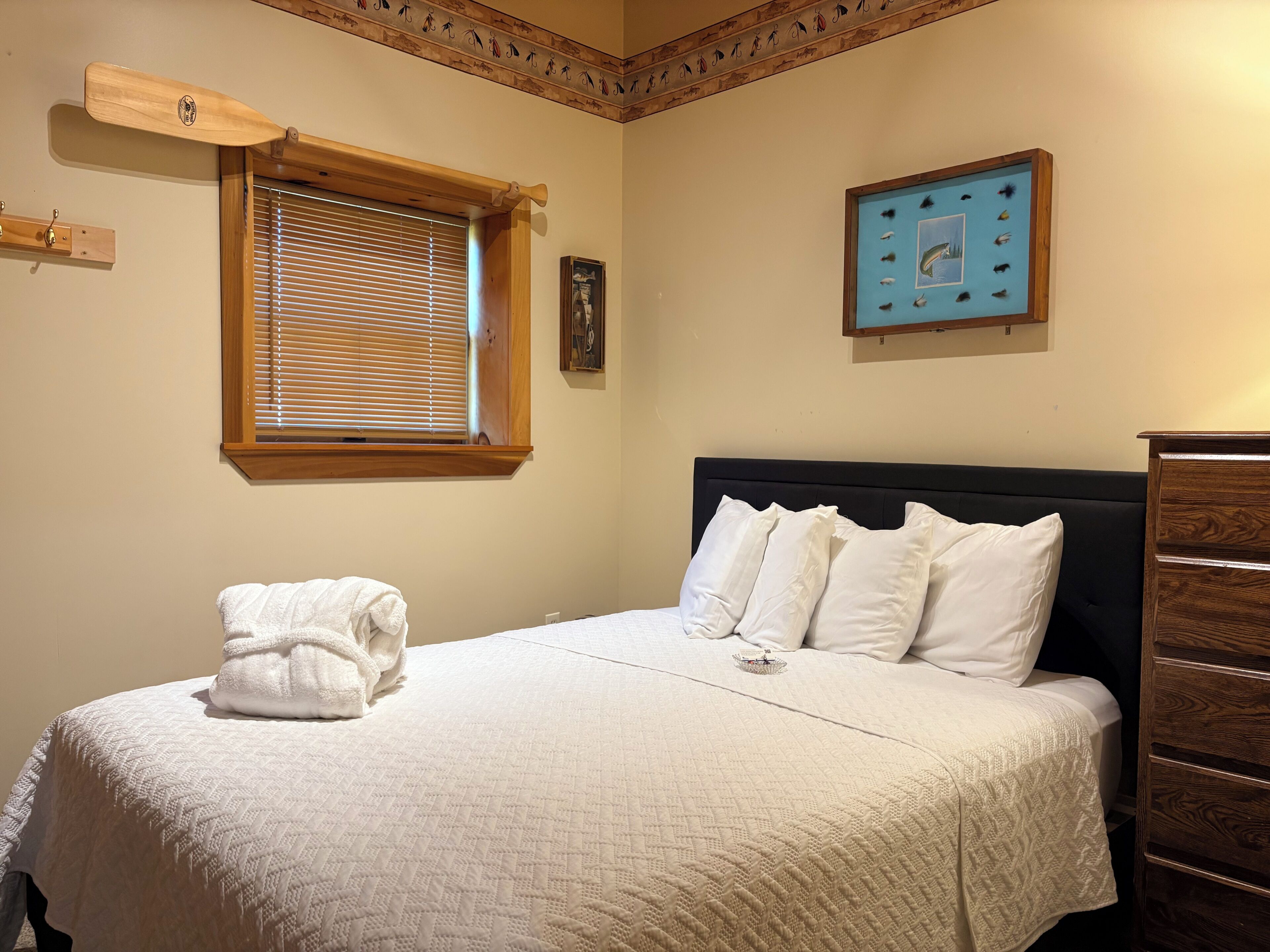 A neatly made bed with a white comforter and pillows, a wooden headboard, a window with blinds, and a framed picture on the wall.