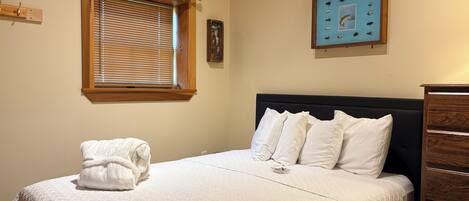 A neatly made bed with a white comforter and pillows, a wooden headboard, a window with blinds, and a framed picture on the wall.