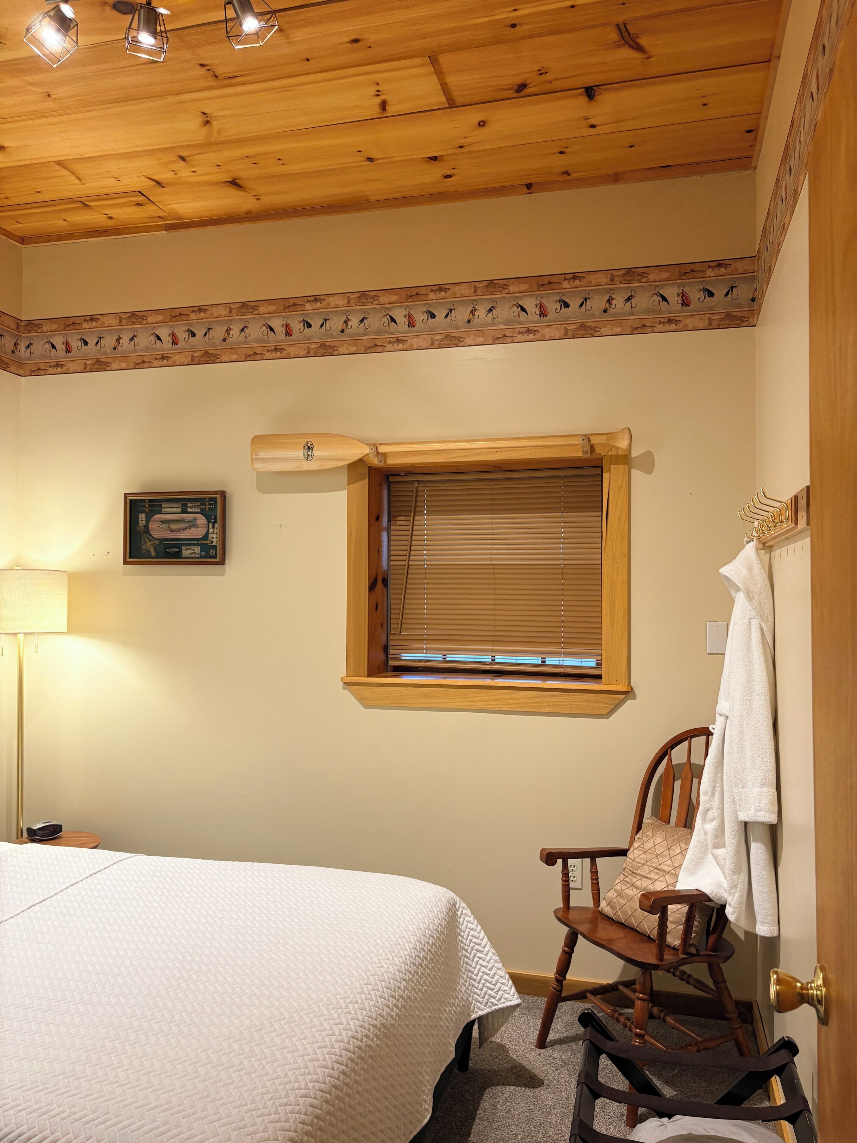 Una camera da letto con soffitto in legno, un letto, una sedia, una finestra con tende e una lampada, oltre a un quadro appeso alla parete.