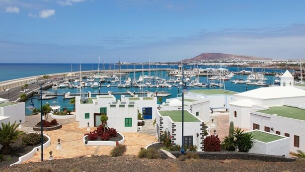 Un puerto deportivo con numerosas embarcaciones amarradas, rodeado de edificios blancos con detalles verdes y una vista despejada del mar y colinas distantes.