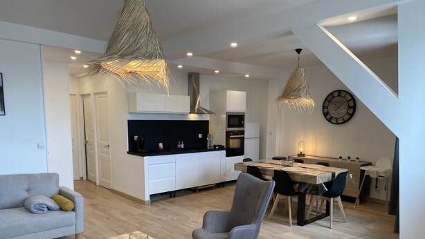 Ein moderner Wohnraum mit Sofa, Couchtisch, Essbereich mit Tisch und Stühlen, Küche mit weißen Schränken und Geräten sowie einer Pendelleuchte.