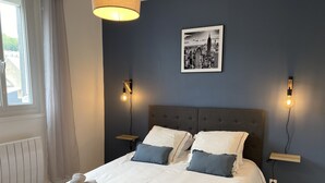 Ein ordentlich bezogenes Bett mit weißer und blauer Bettwäsche, zwei Nachttische mit Lampen, ein Stadtbild in einem Bilderrahmen und ein Fenster mit Vorhängen.