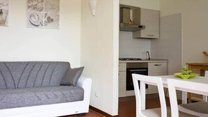 A modern kitchen with white cabinets, a stove, and a sink. There is a grey sofa and a white dining table with chairs.