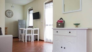 A living room with a white sofa, a dining table with chairs, a white cabinet, a television, and a clock on the wall.