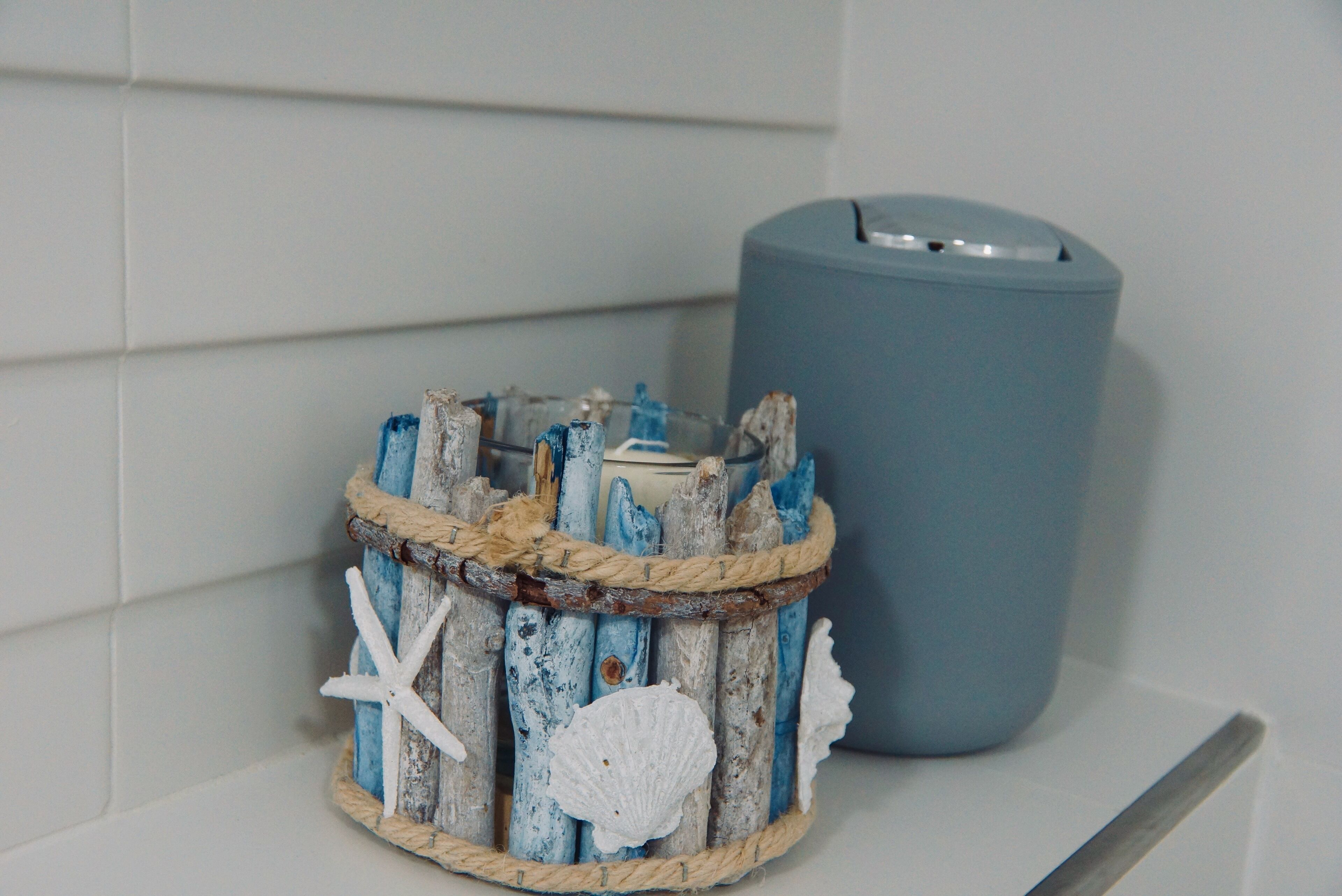A rustic wooden basket with seashells and a rope handle, placed next to a blue trash can.