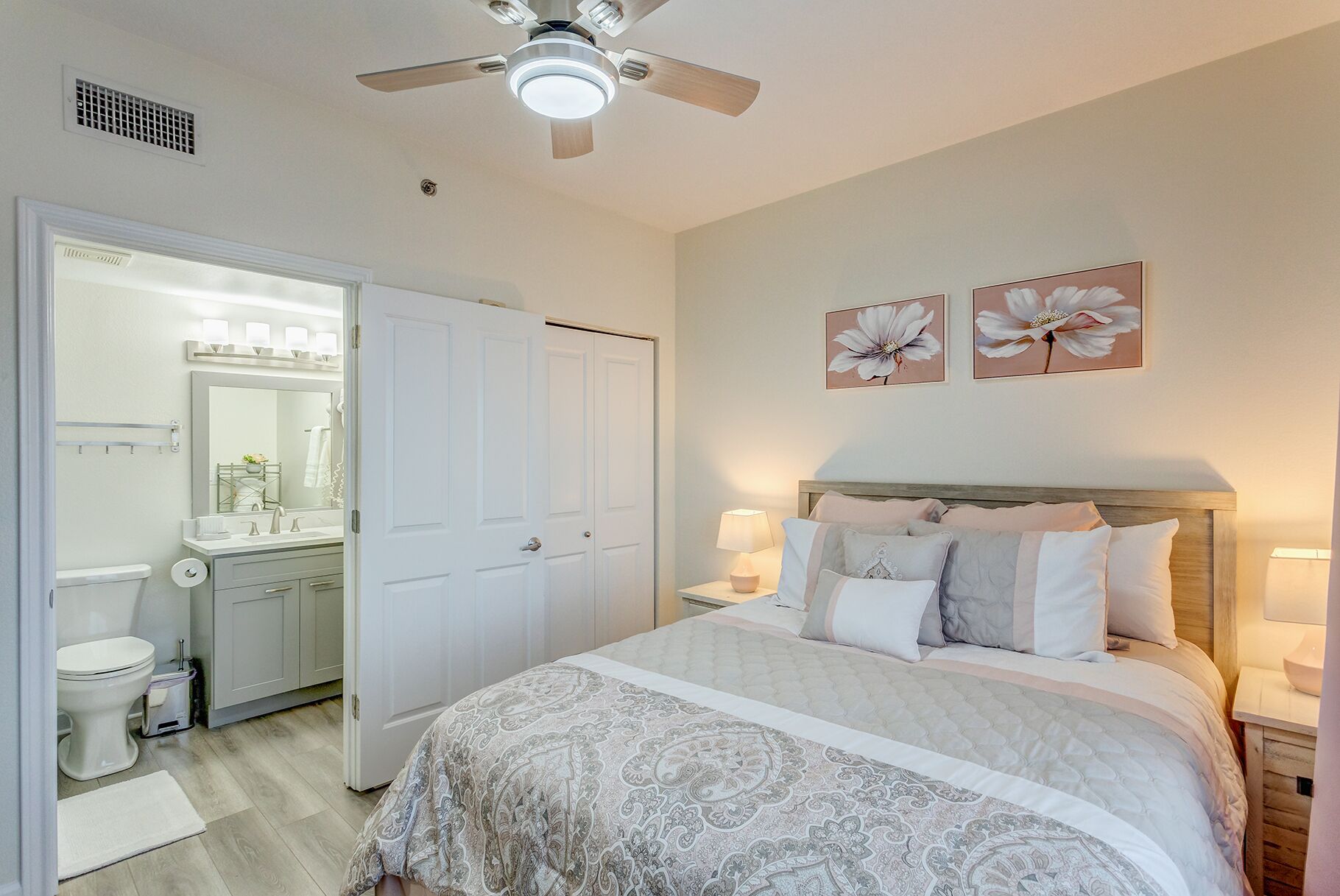 A bedroom with a bed, bedside lamps, a ceiling fan, and two wall-mounted flower paintings.