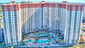 A high-rise hotel with multiple balconies, a central pool area with palm trees, and a clear view of the surrounding landscape.