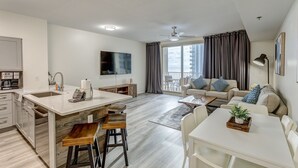 A modern apartment with a kitchen, dining area, and living room. It includes a large window, a ceiling fan, and a flat-screen TV.