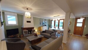 Amplia sala de estar con un sofá grande de color gris, una mesa de centro de madera y una chimenea. Hay varias sillas y una mesa de comedor con sillas. El piso es de madera y hay ventanas grandes.