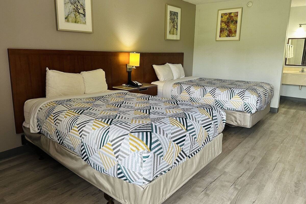 A hotel room with two beds, a wooden headboard, a nightstand with a lamp, and a bathroom with a sink and mirror.