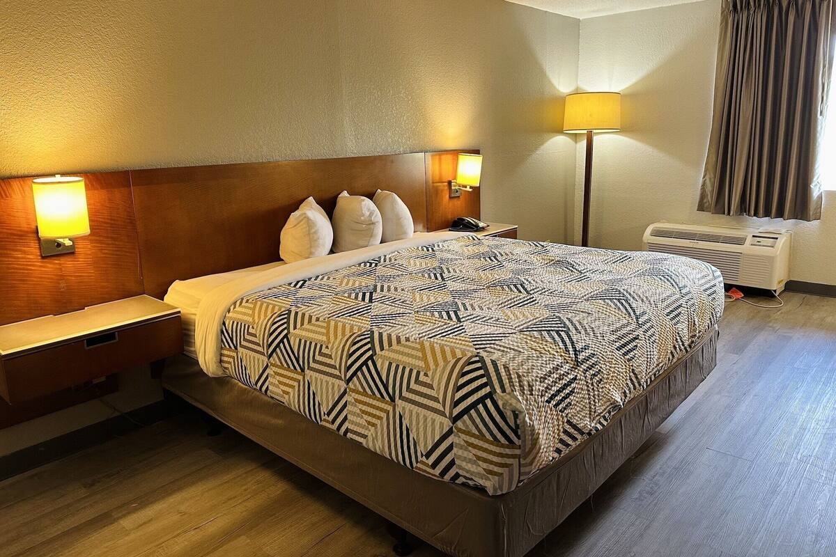 A hotel room with a large bed, two bedside tables, a nightstand, a lamp, an air conditioning unit, and a window with curtains.