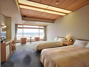 A hotel room with a large window, two beds, a desk with a chair, a small table, and a nightstand with a lamp. - Hotel Urashima (Nachikatsuura)
