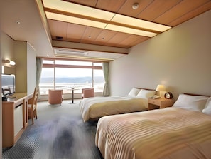 A hotel room with a large window, two beds, a desk with a chair, a small table, and a nightstand with a lamp.