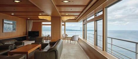 A spacious deck area with a sofa, chairs, a table, and large windows offering a view of the sea.