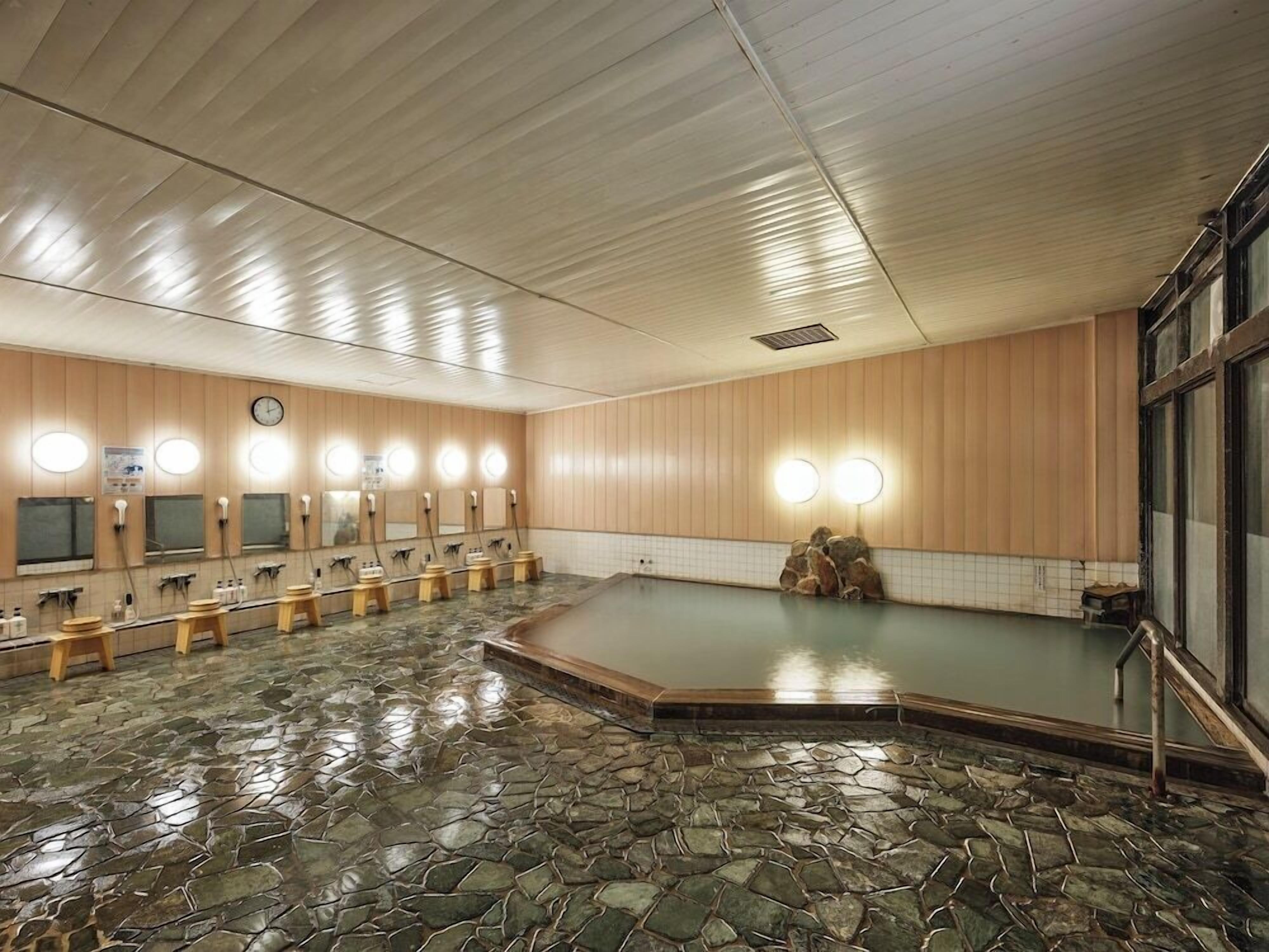 A public bath area with multiple washbasins, a stone-tiled floor, wooden panel walls, and a large window on the right.