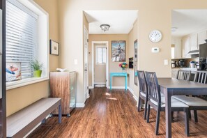 A hallway with a wooden floor, a bench, a table with chairs, a clock, and a window with blinds.