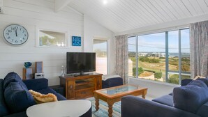 A living room with a television, sofas, a coffee table, and a large window offering a view of the beach. - Wave and See - 53 Gold Coast Drive (Carrickalinga)