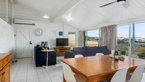 A modern living room with a wooden dining table, blue sofas, and a TV. - Wave and See - 53 Gold Coast Drive (Carrickalinga)