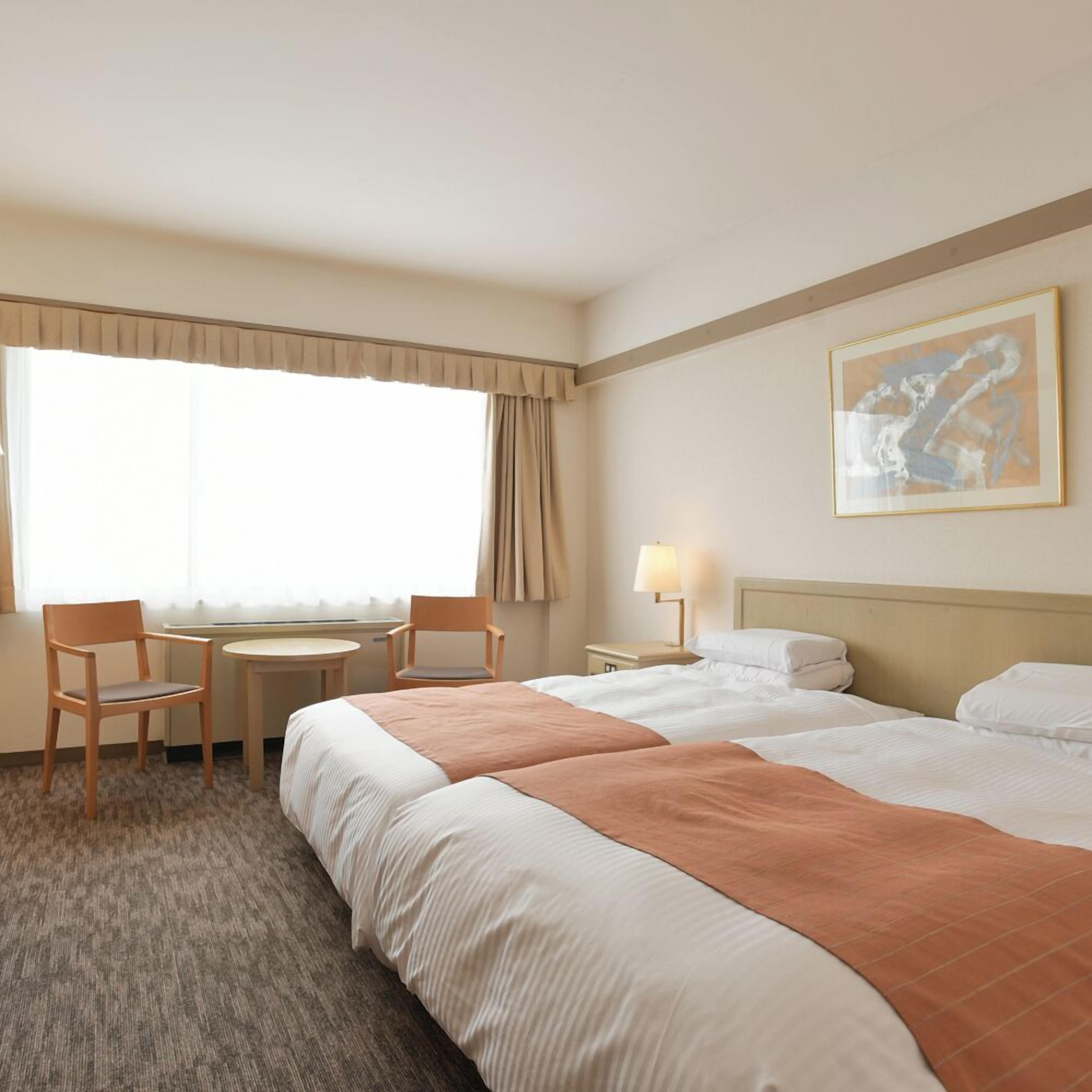A hotel room with two beds, a desk, two chairs, a lamp, and a painting on the wall.