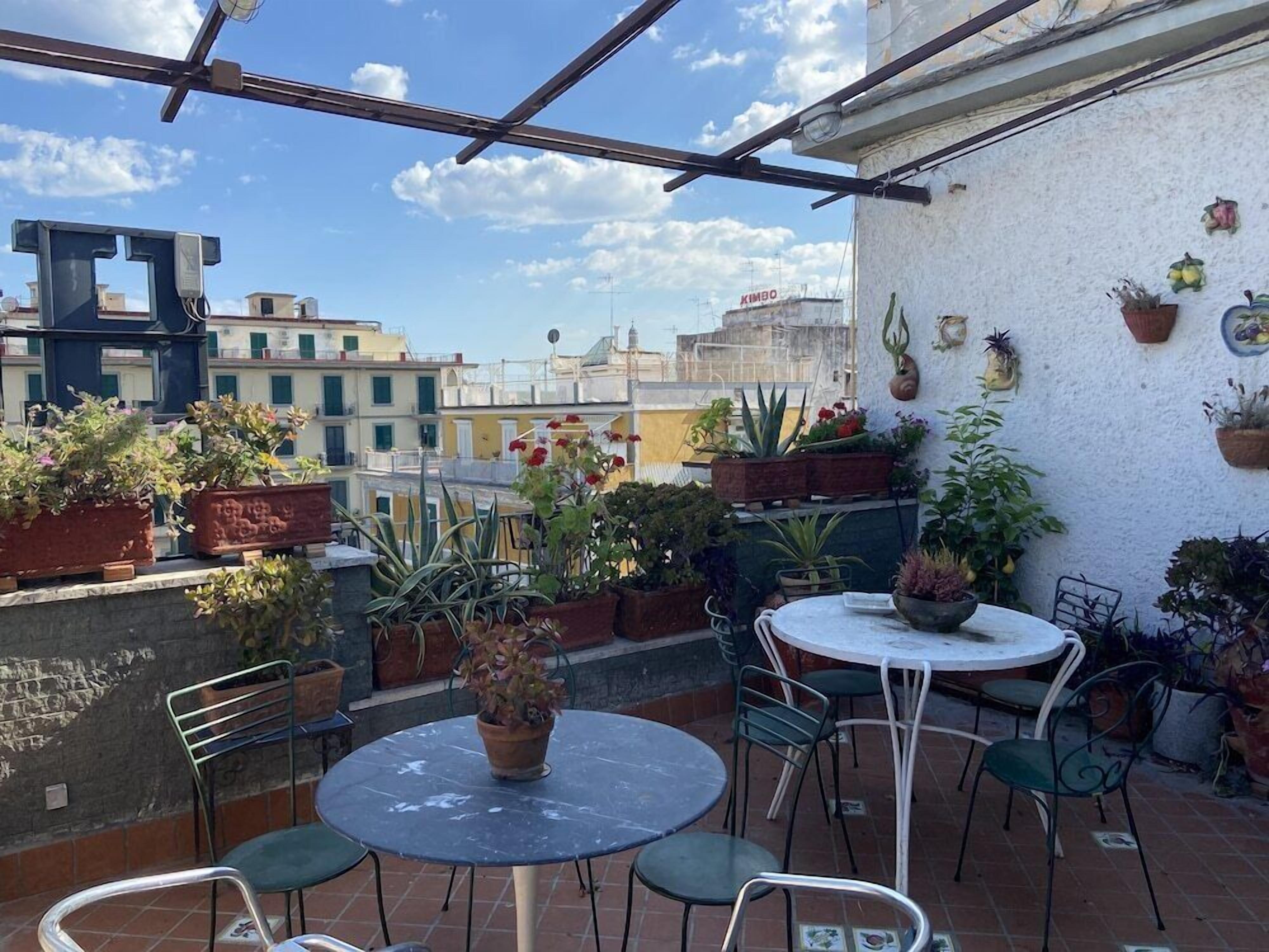 A rooftop terrace with potted plants, a round table, and chairs.