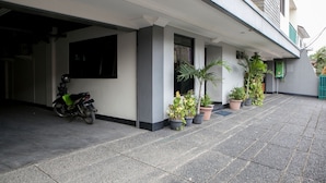 A covered outdoor area with a motorcycle parked to the left, potted plants along the walkway, and a building entrance with a green door. - Reddoorz @ Buah Batu (Bandung)