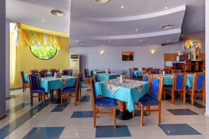 A restaurant with multiple tables and chairs, a large window with curtains, and a kitchen area visible in the background. - Evridika (Nessebar)