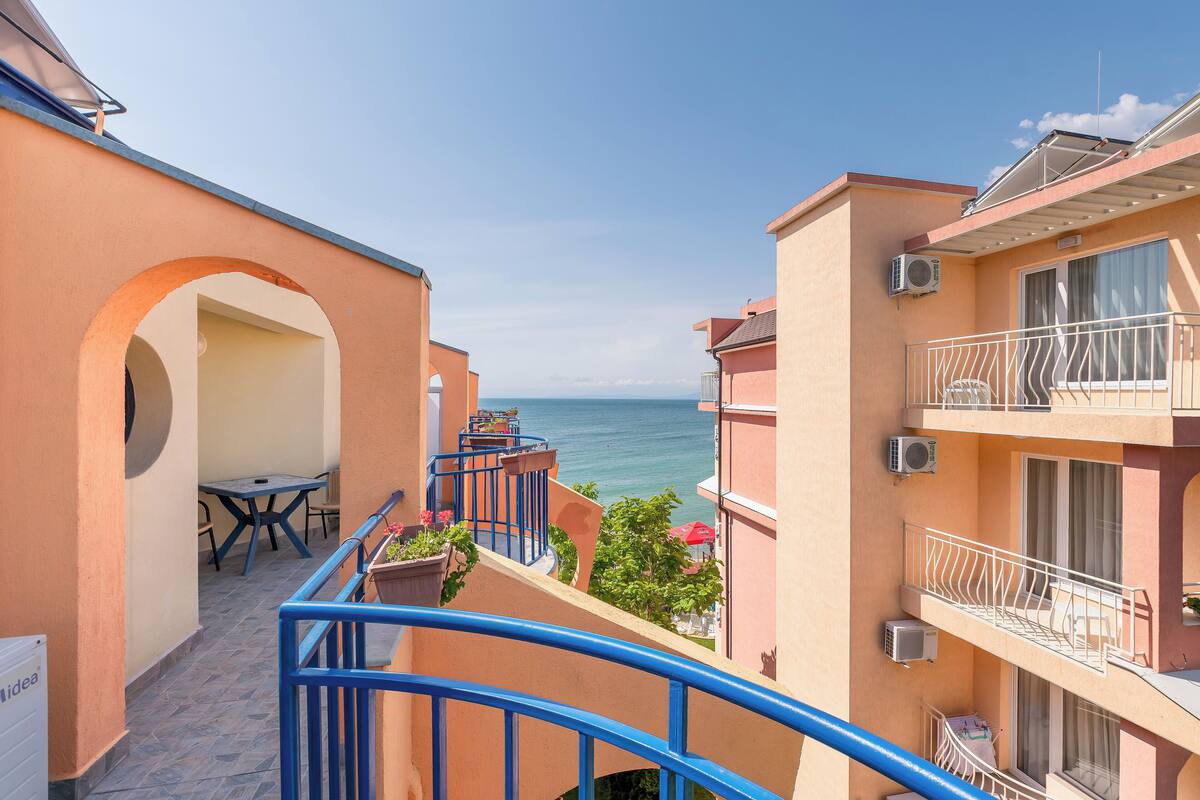 A balcony with a view of the sea, a table with chairs, and a small garden area.