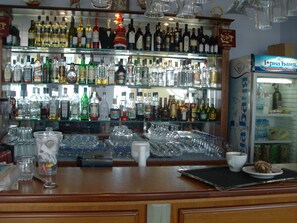 Zona de bar con estantes de diversas botellas de licores, vasos y un refrigerador con una etiqueta.