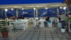 An outdoor dining area with white tables and chairs, a stage with a microphone stand, and potted plants. - Evridika (Nessebar)