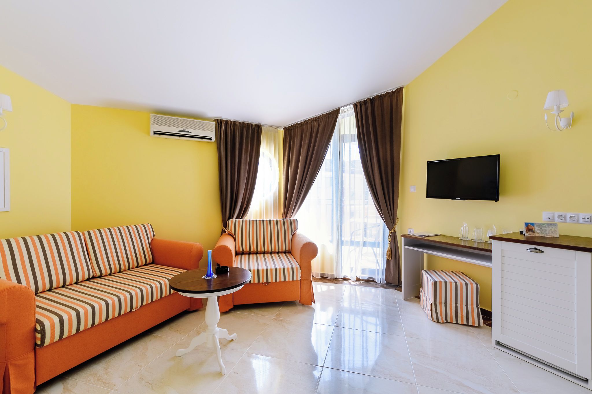 A room with a sofa, armchair, small table, TV, and a kitchenette area.