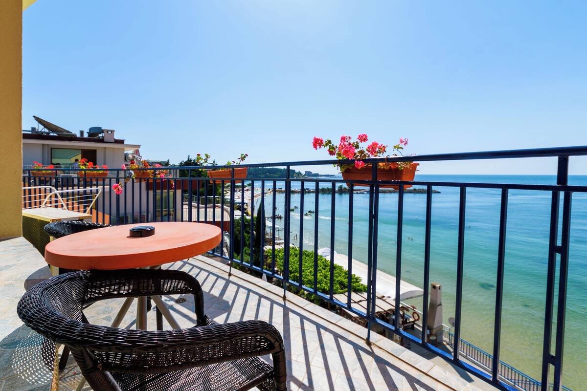 A balcony with a round table and chairs, potted plants, and a view of the sea.