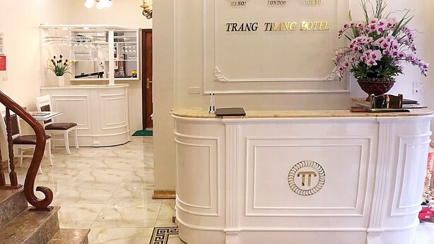 A hotel reception area with a marble floor, a reception desk with a logo, a clock displaying time in three cities, and a staircase leading to another room. - Trang Trang Premium Hotel (Hanoi)