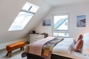 Une chambre avec un plafond en pente, un puits de lumière, un lit recouvert d’une courtepointe, un banc, une table de chevet et deux tableaux accrochés au mur.