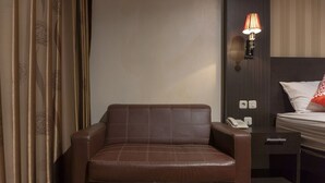 Une chambre d’hôtel avec un fauteuil en cuir marron, une table de chevet avec une lampe, un téléphone et un lit avec un oreiller blanc et un coussin décoratif rouge.