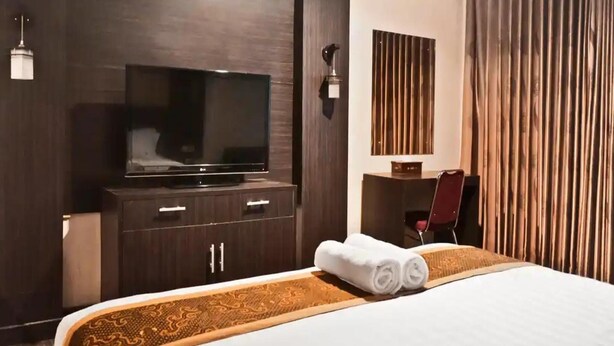 Une chambre d’hôtel équipée d’un grand téléviseur à écran plat, d’une commode en bois avec des tiroirs, d’un bureau avec une chaise et d’un lit sur lequel est posée une serviette enroulée.
