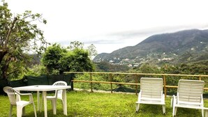 A grassy outdoor area with a table and chairs, a wooden fence, and a view of a valley with buildings and hills.