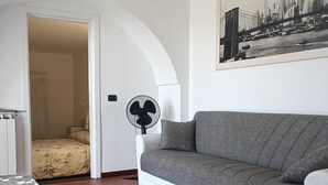 A living room with a sofa, a fan, a tiled floor, and a door leading to another room.